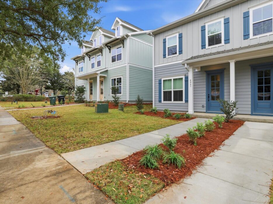 Two modern townhouses with blue accents, tidy landscaping, and a concrete sidewalk leading to blue front doors. Trees and a green lawn line the residential street under a blue sky.