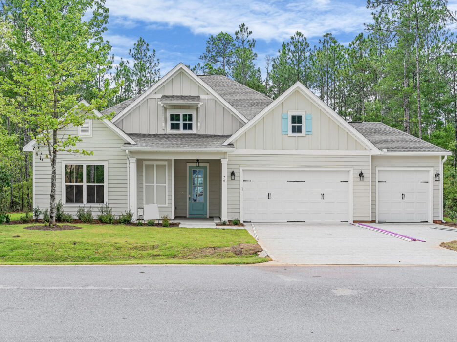 Single-story suburban house with white siding, a covered front porch, and a double garage. The lawn is freshly mowed and surrounded by trees and greenery, with a clear sky and paved driveway out front.