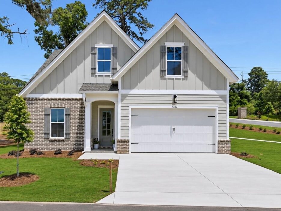 A modern suburban house with gray siding, brick accents, two front gables, white garage door, manicured lawn, new landscaping, and a clear blue sky in the background.